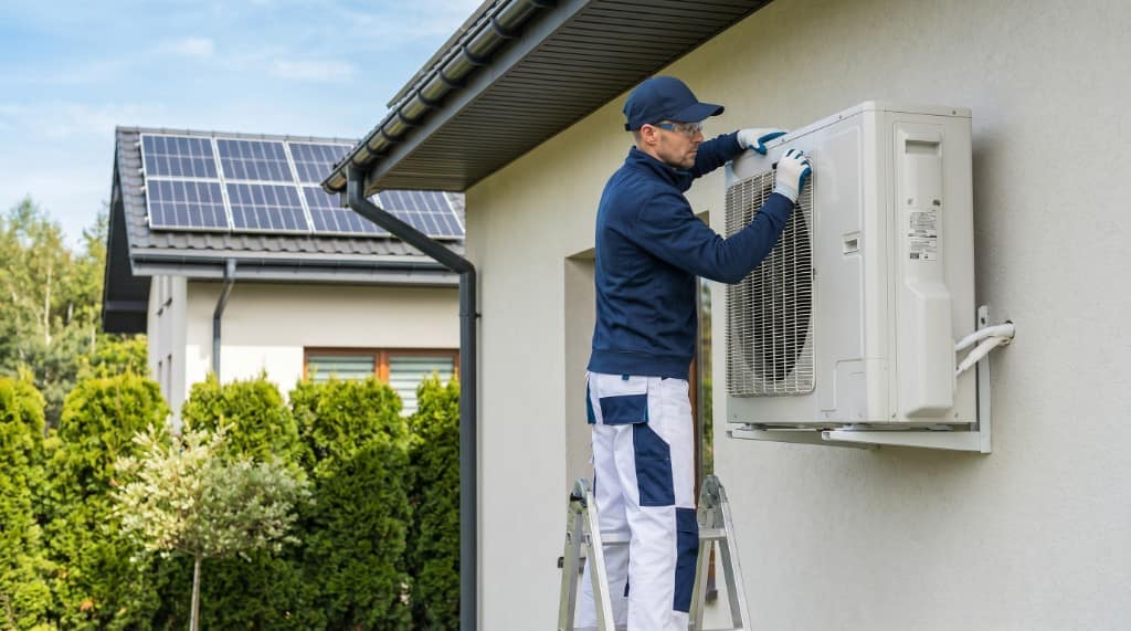 Technician on a ladder installing an outdoor heat pump unit on the exterior wall of a New Zealand home with solar panels on the roof.